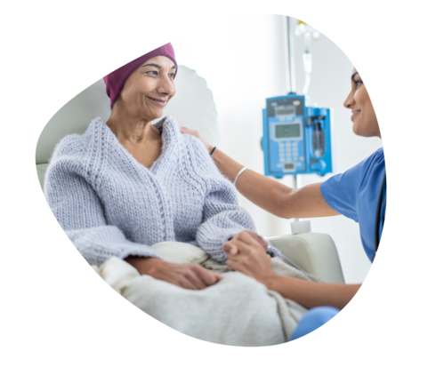 Eine Frau sitzt auf einem Behandlungsstuhl und bekommt eine Infusion. Eine andere Frau hält ihre rechte Hand und guckt freundlich.