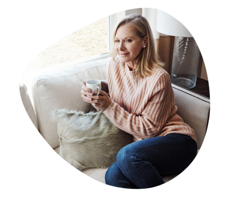 Eine Frau mit einem dicken Pullover und einer Tasse in beiden Händen sitzt auf einem Sofa und schaut zuversichtlich.