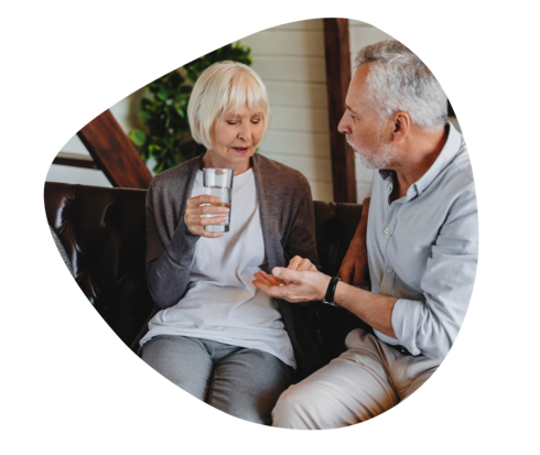 Ein Mann hält einer Frau in seiner offenen linken Hand eine Tablette hin. Sie hat in der rechten Hand ein Glas Wasser, mit der linken greift sie zur Tablette.
