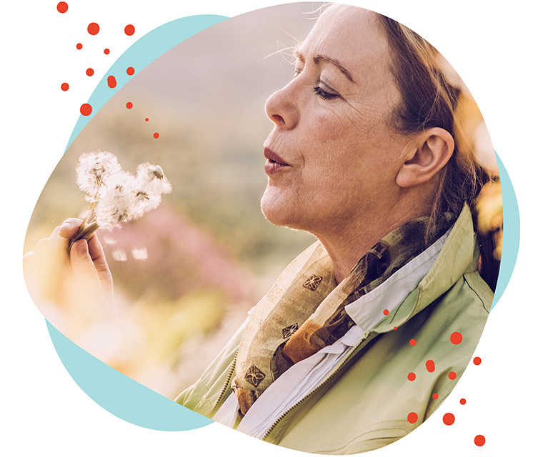 Leben mit Brustkrebs: Eine brünette Frau pustet gegen eine Pusteblume in ihrer Hand