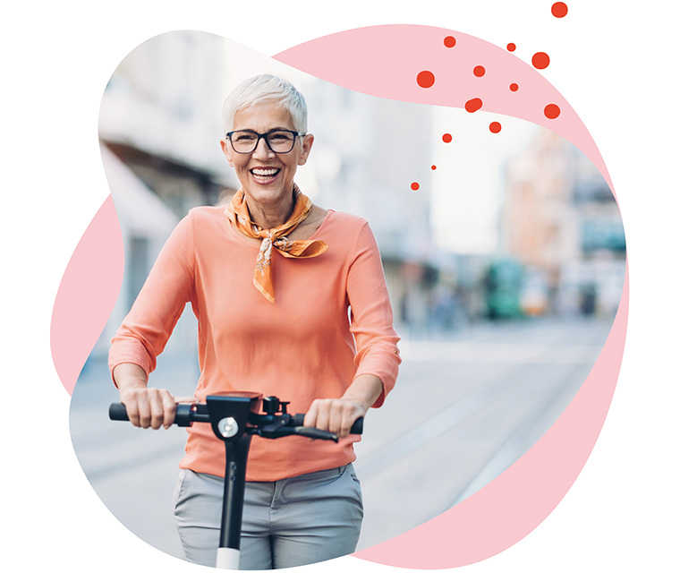 Leben mit Brustkrebs: Eine grauhaarige Frau fährt lachend mit einem Elektroroller Leben mit Brustkrebs: Eine grauhaarige Frau fährt lachend mit einem Elektroroller