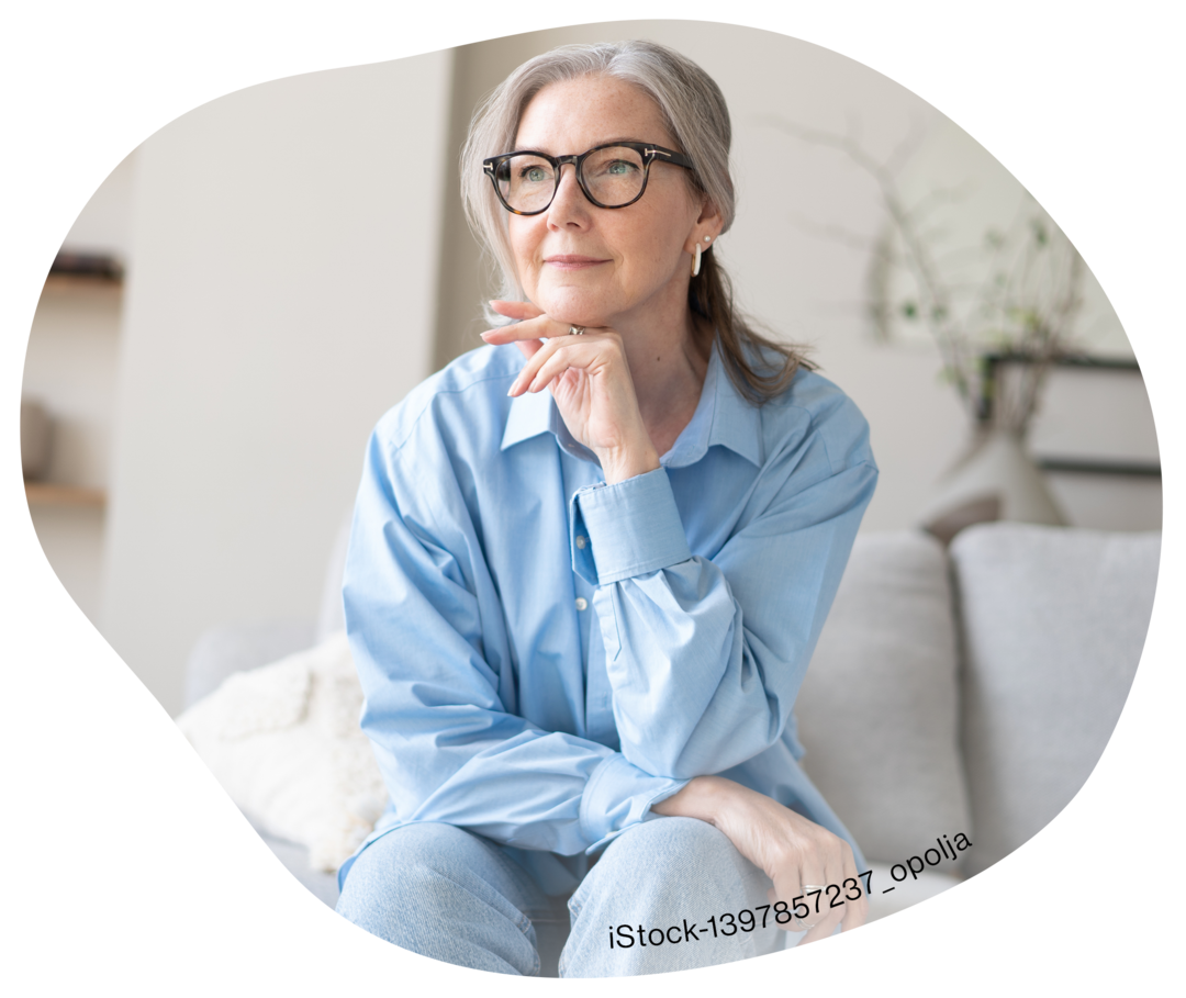 Eine ältere Frau mit Brille sitzt nachdenklich auf einer Couch.