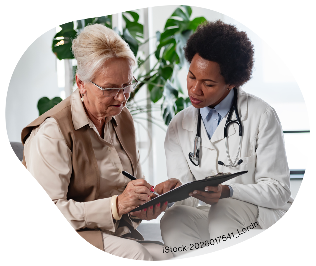 Eine ältere Frau schaut zusammen mit einer Frau im weißen Kittel auf ein Klemmbrett.